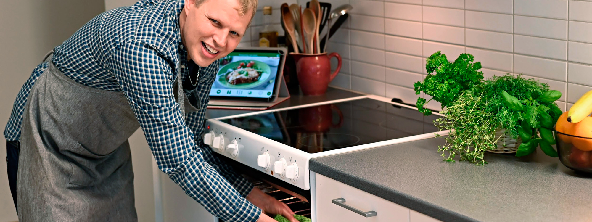 En man tar ut en form ur en ugn. En surfplatta med appen MatGlad 2.0 står på köksbänken.