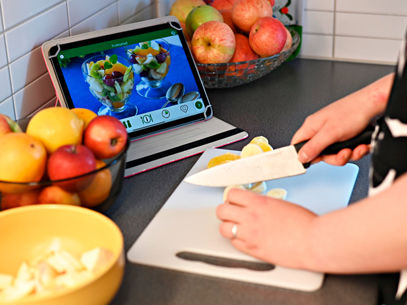 Händer som skär frukt till fruktsallad. MatGlad 2.0 i bakgrunden